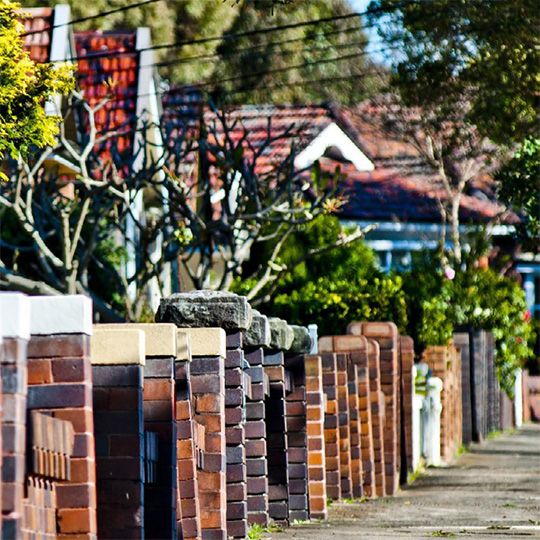 A view down an inner suburban footpath of a row of brick fences and early-20th-century single-family housing.